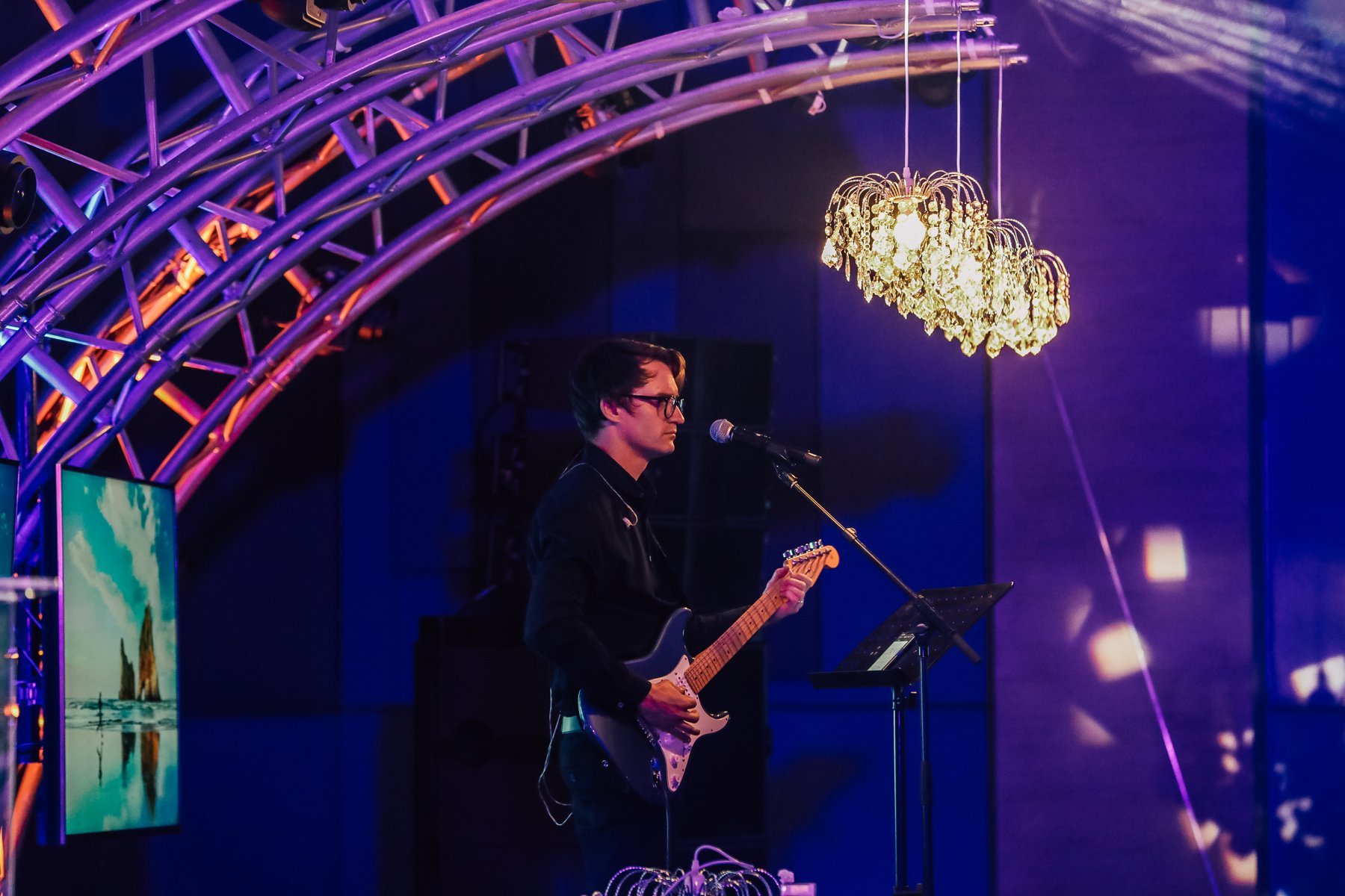 Electric guitarist at a microphone under a crystal chandelier and magenta-lit truss