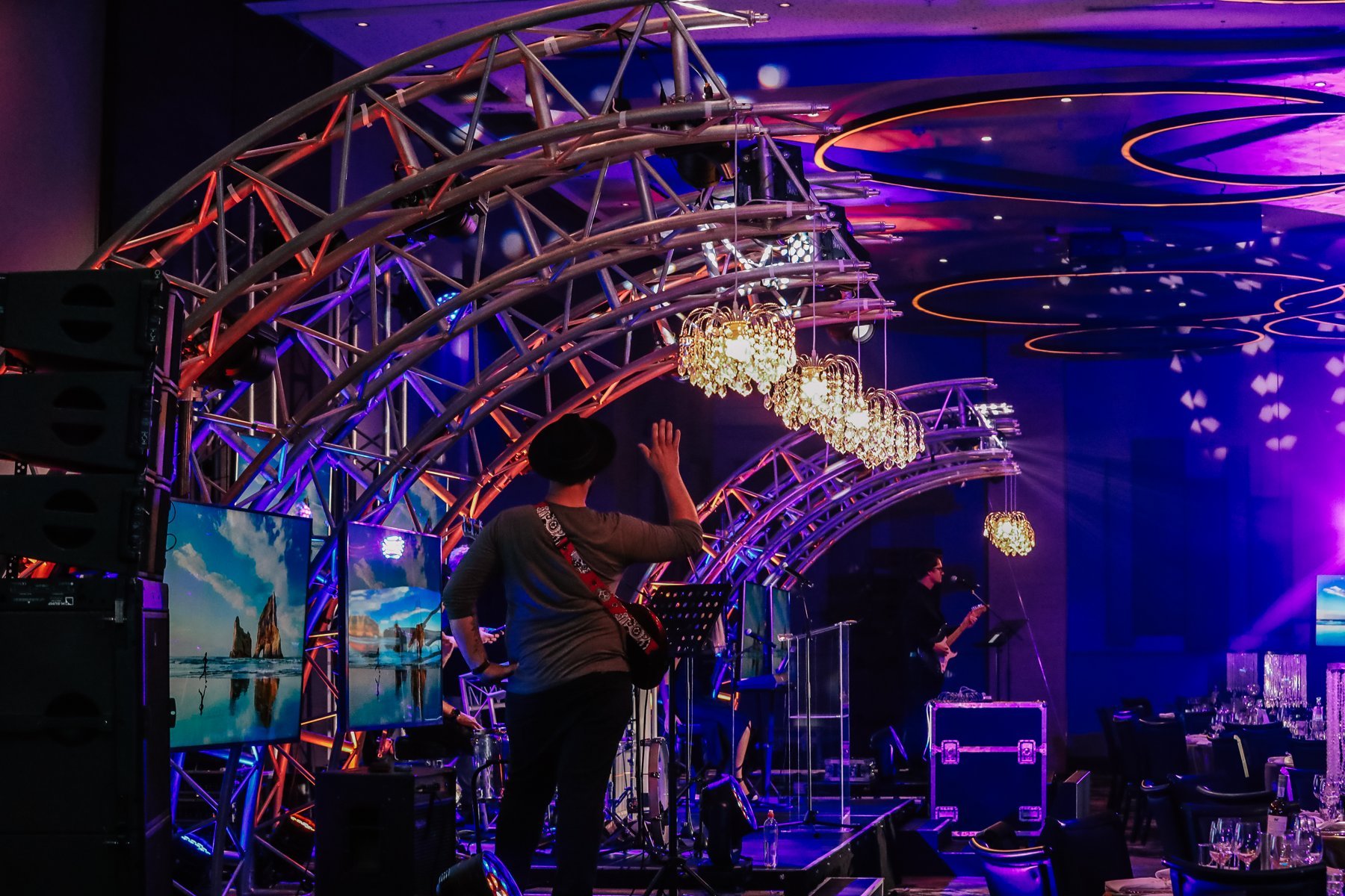 Stage under curved silver trusses with crystal chandeliers, a musician, and coastal landscape monitors