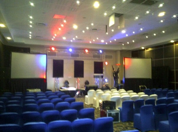 Setup state with blue chair covers and a white VIP block facing dual blank screens under red and blue truss spots, with flight cases on the floor