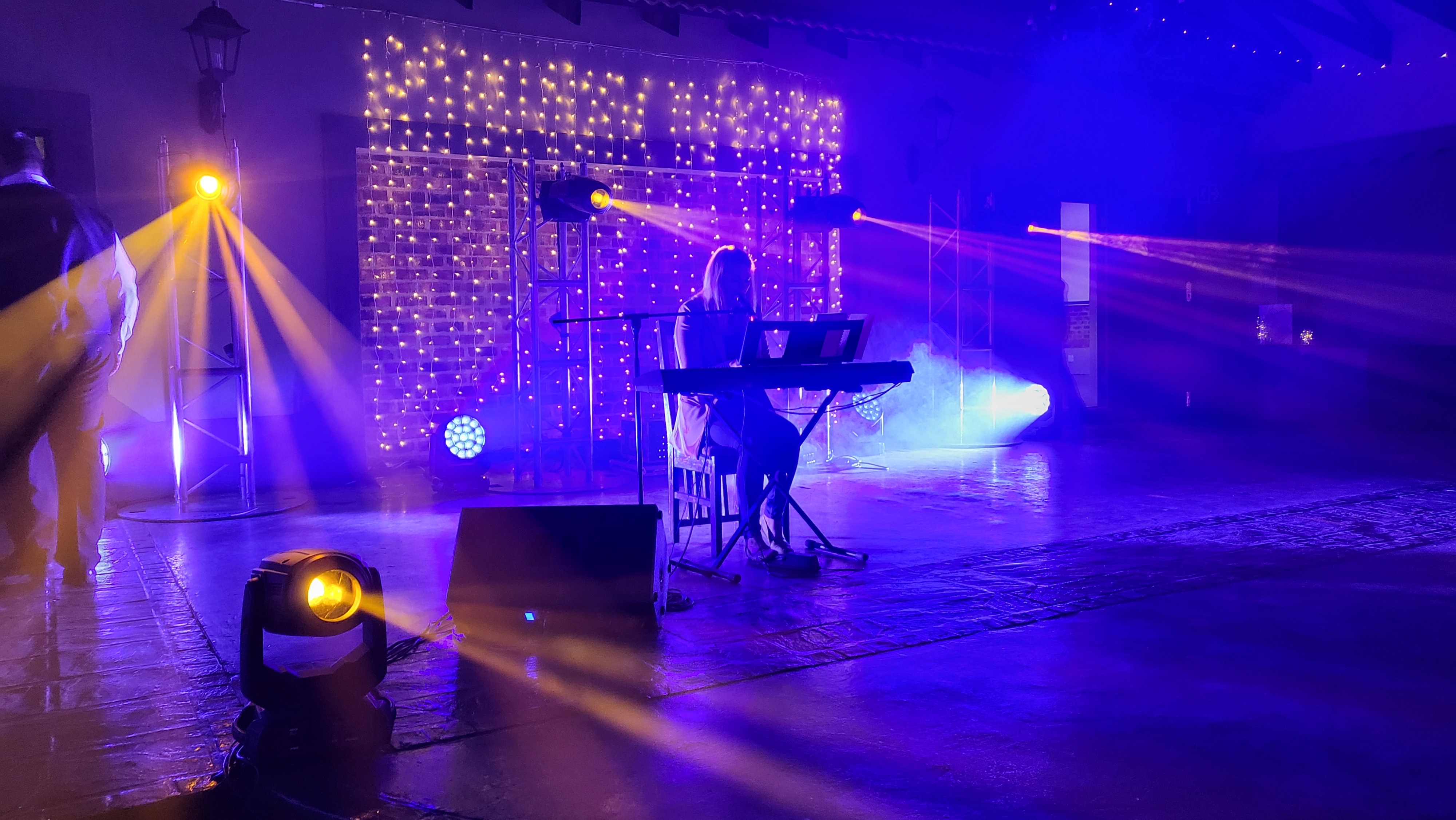 Keyboard player on stage in front of a fairy-light curtain with haze, truss towers, and yellow and purple beams