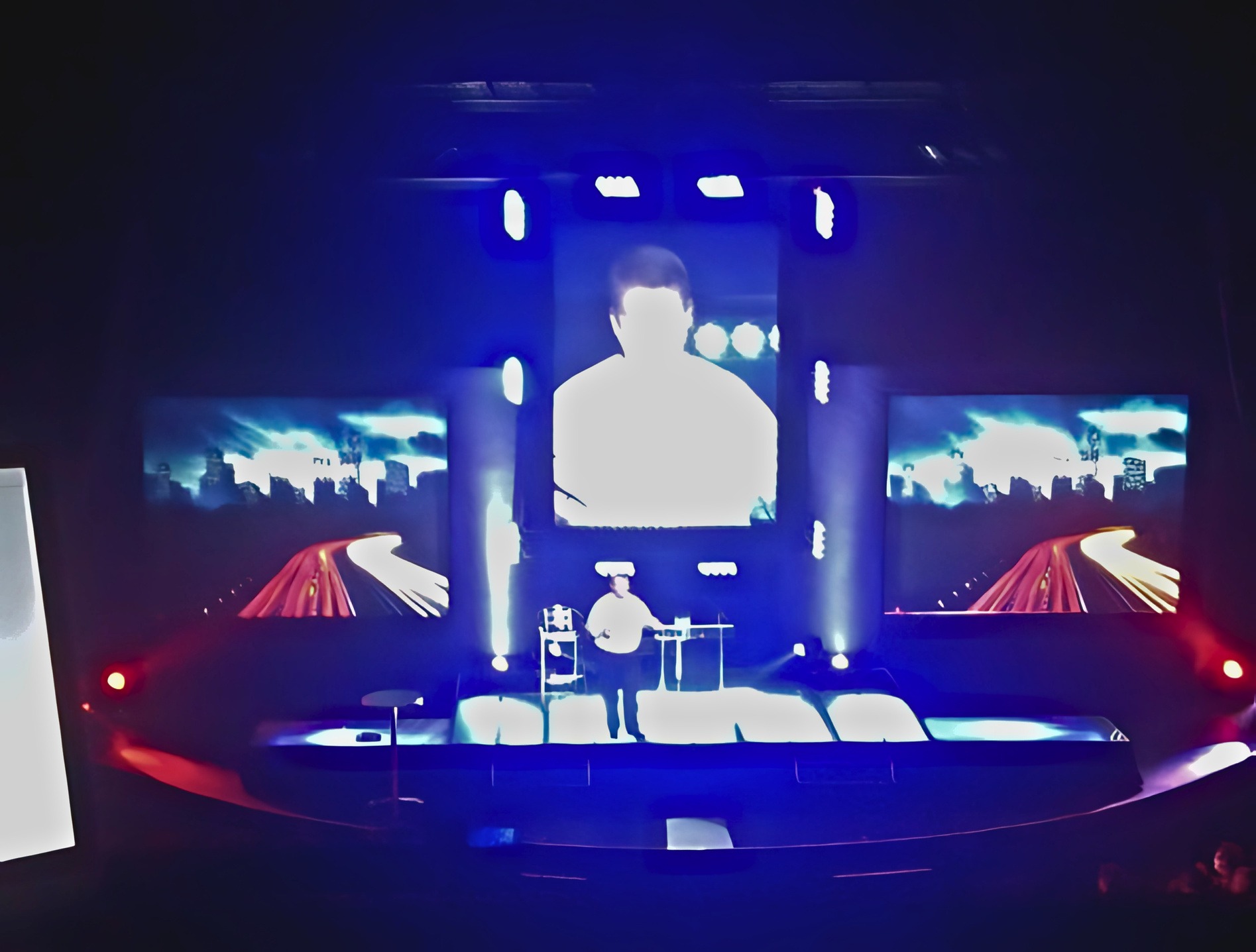 Live keynote with the speaker on centre IMAG between dual side LEDs showing city night-trails under a blue and white stage wash