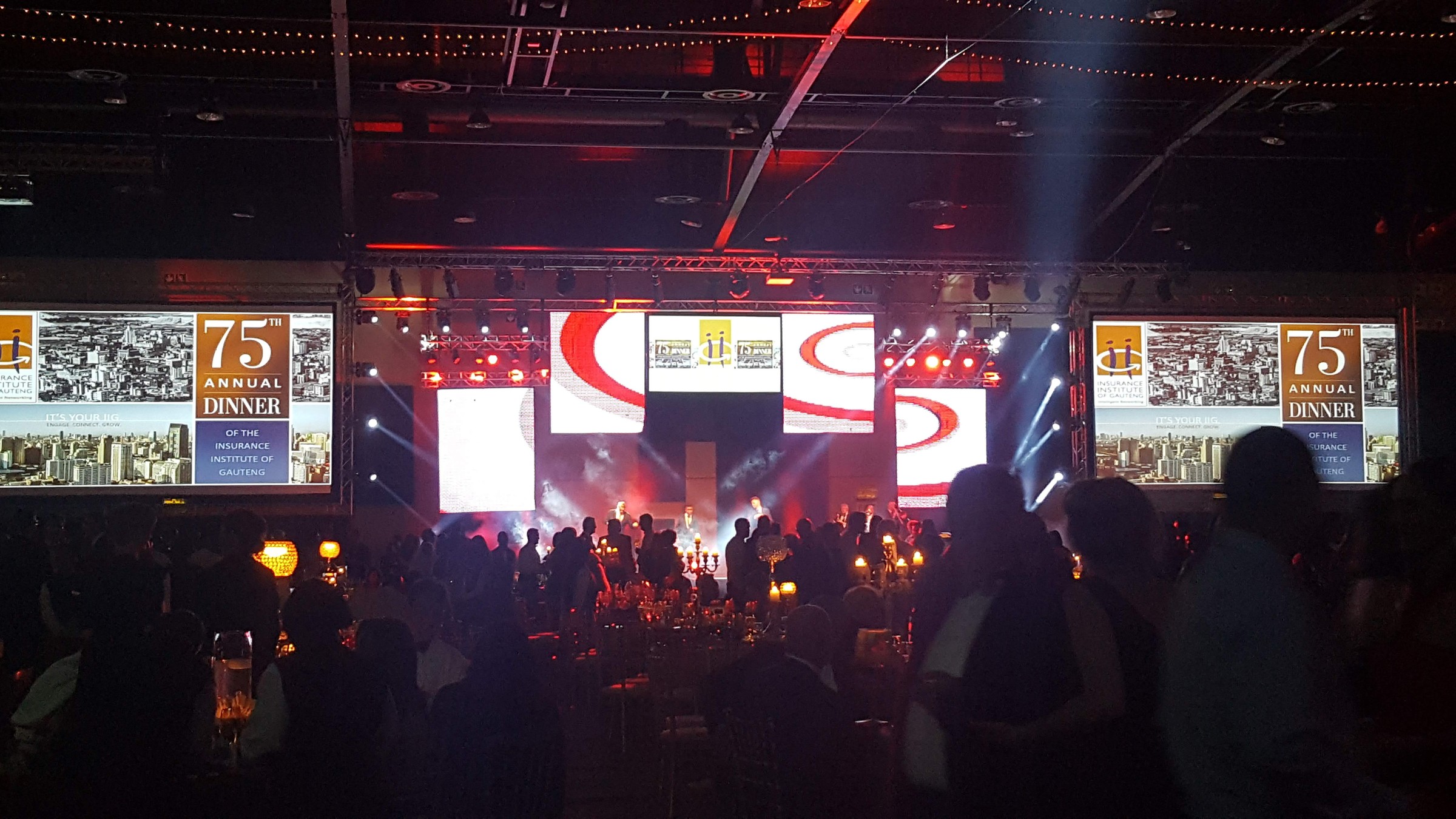 Stage haze with white and blue beams, multi-panel LED cluster, and IIG 75th Annual Dinner side screens