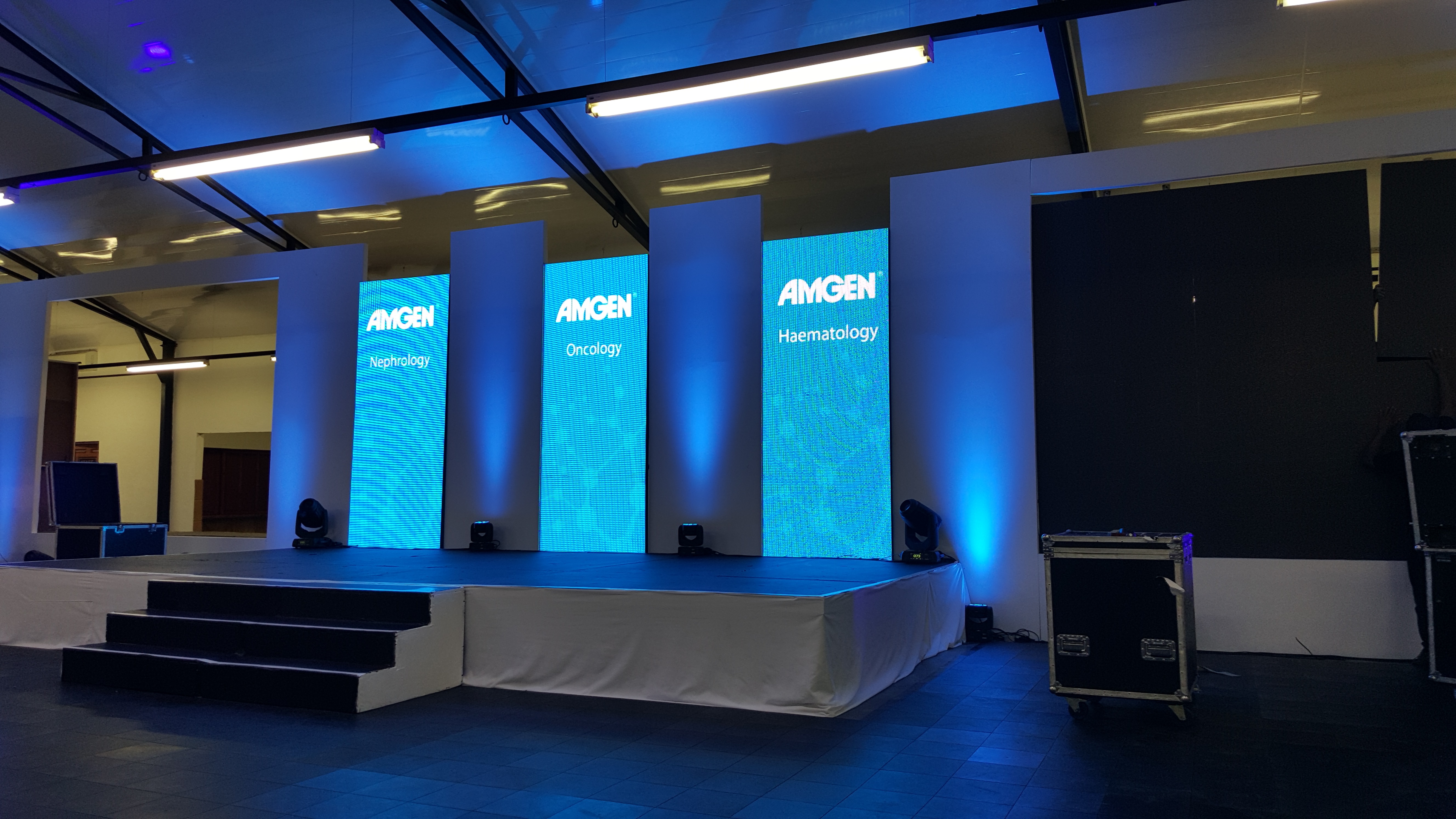 Stage with three vertical LED panels reading AMGEN Nephrology, AMGEN Oncology, and AMGEN Haematology, blue uplights, and a dressed stage