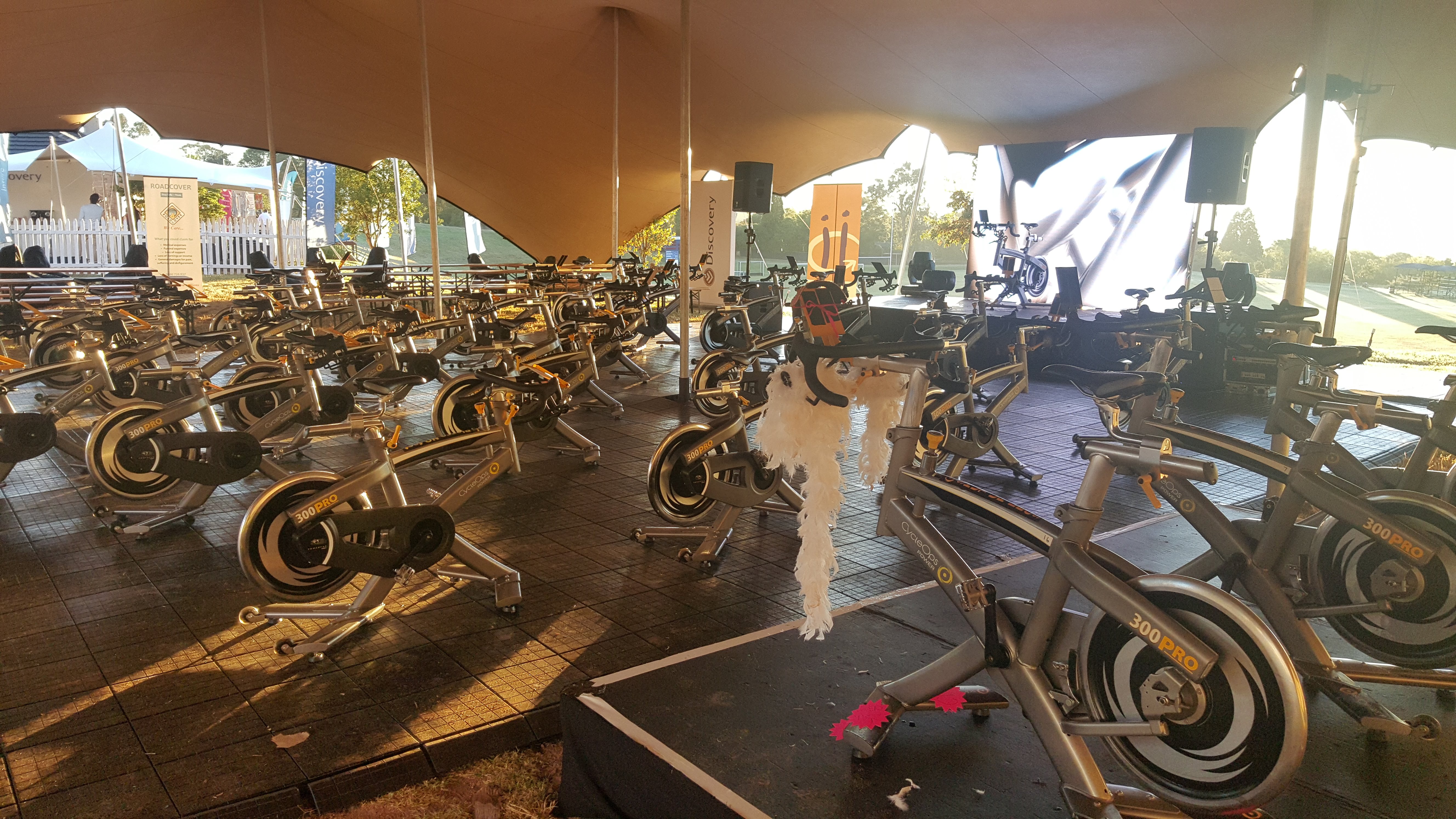 Tan stretch tent interior with rows of spin bikes, a white-feather draped bike, and Discovery and Roadcover banners
