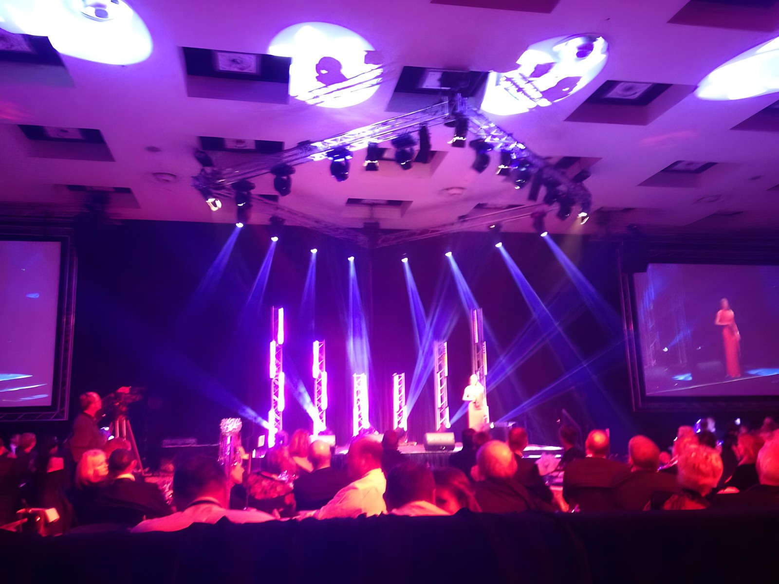 Live floor with purple vertical truss pillars, blue beams, an IMAG screen, a camera operator, and the seated audience