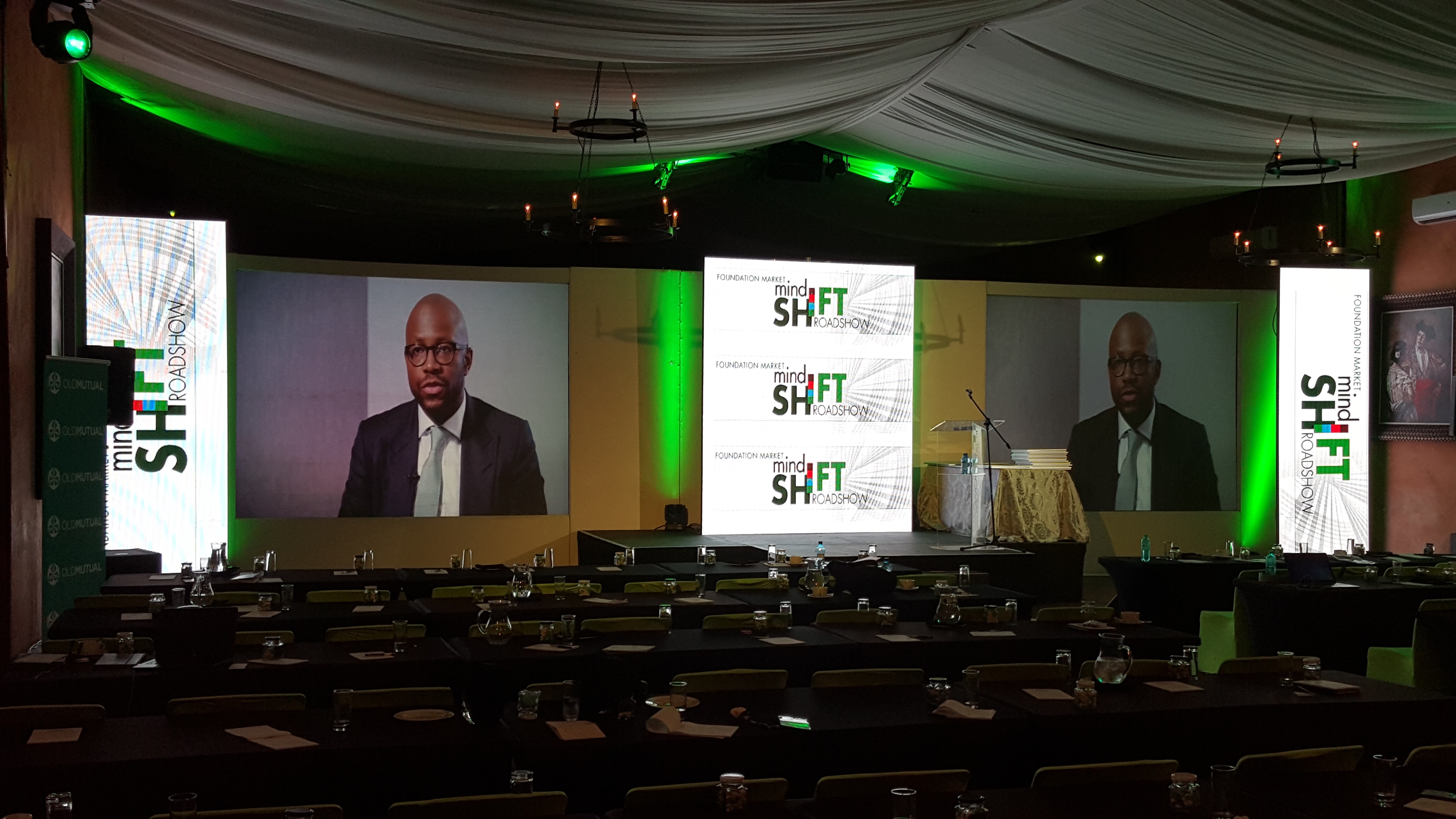 Live speaker shown on dual horizontal screens between mindSHIFT ROADSHOW vertical LEDs, with a green OLD MUTUAL banner, wrought-iron chandeliers, and a green wash