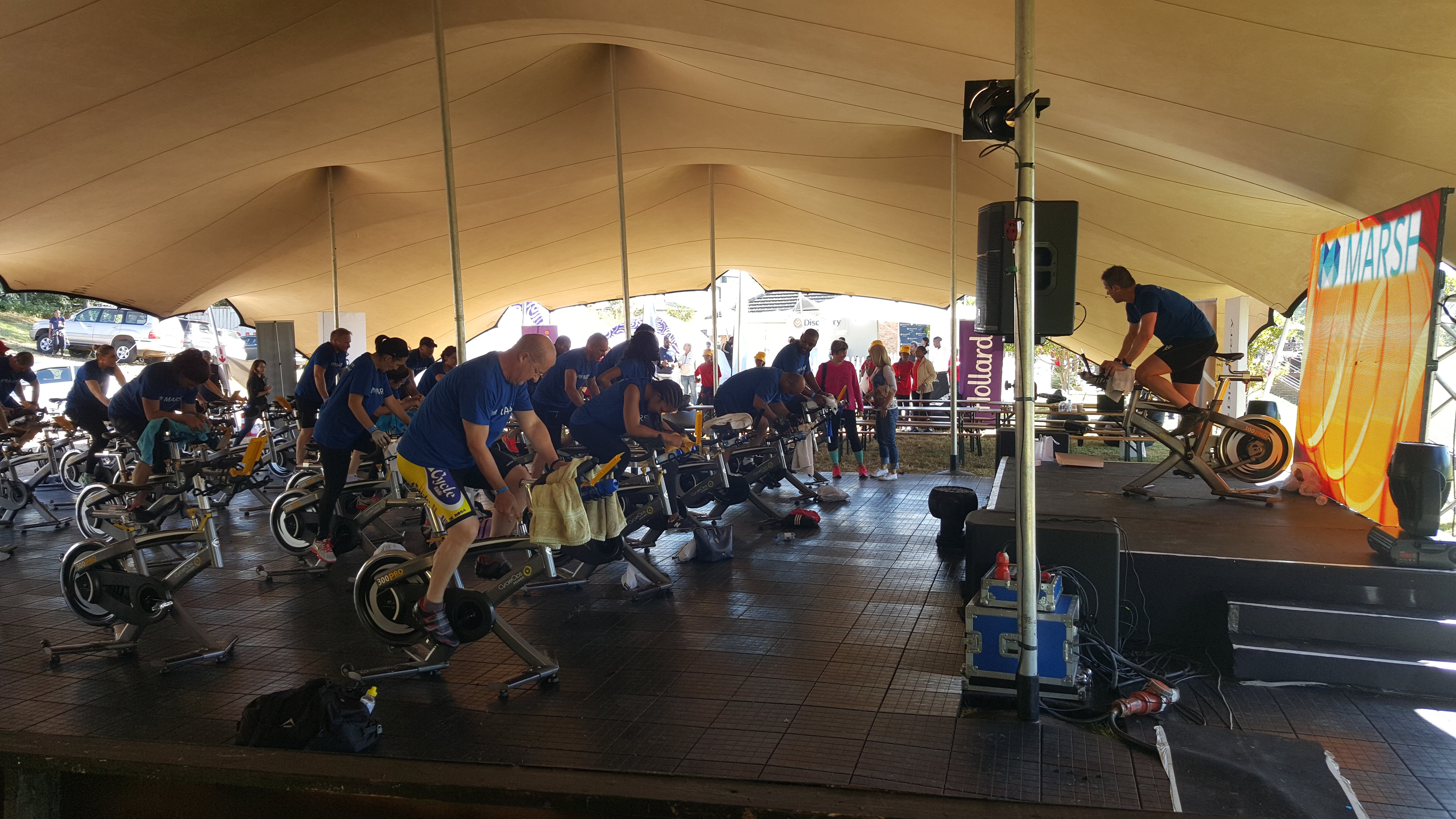 Riders on spin bikes facing a raised stage where an instructor rides beside a stage LED screen reading MARSH, under a stretch tent with a Hollard banner