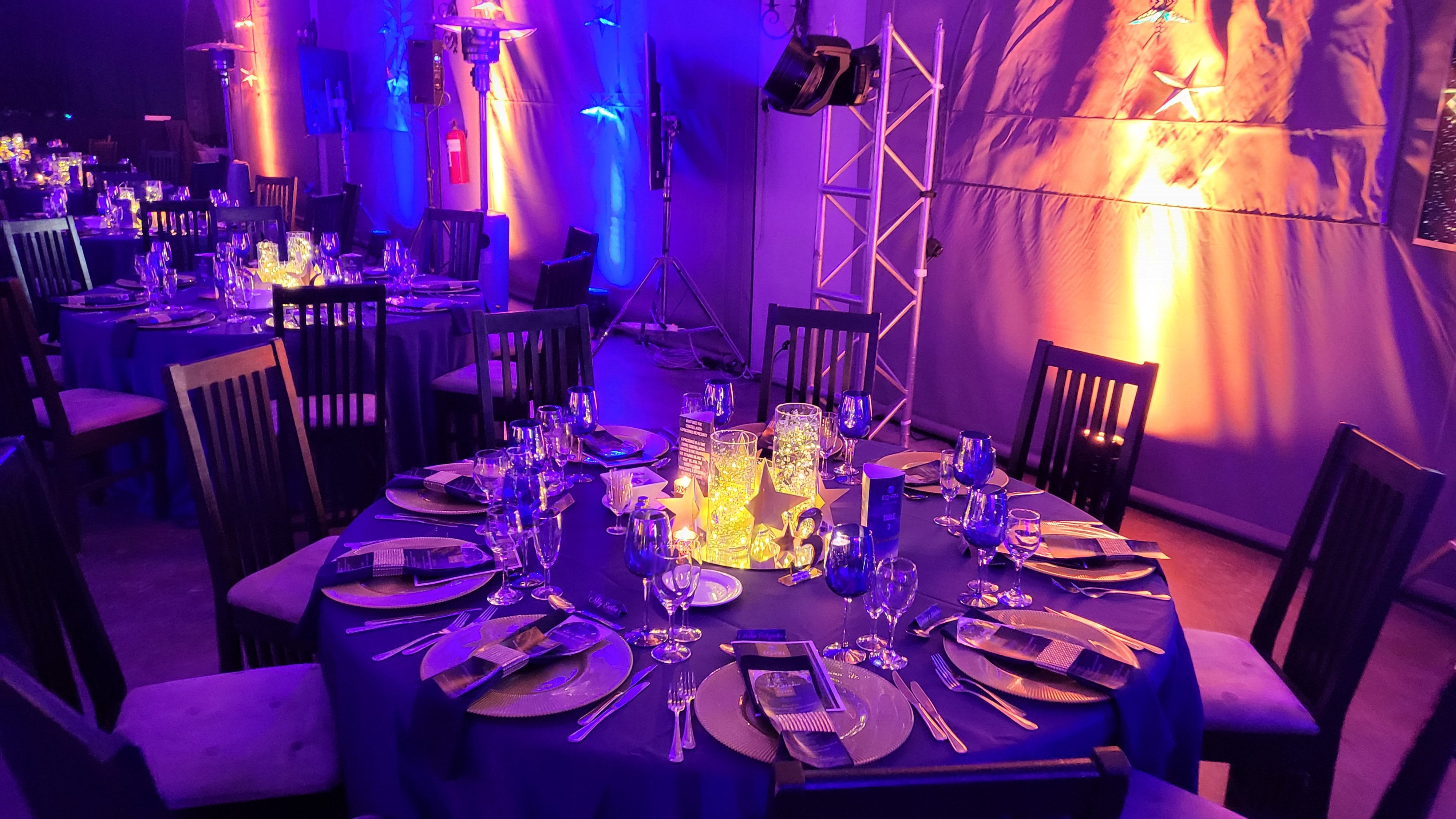 Wide banquet floor with dark tables, gold-star centrepieces, blue and amber wall wash, star gobos, and visible truss