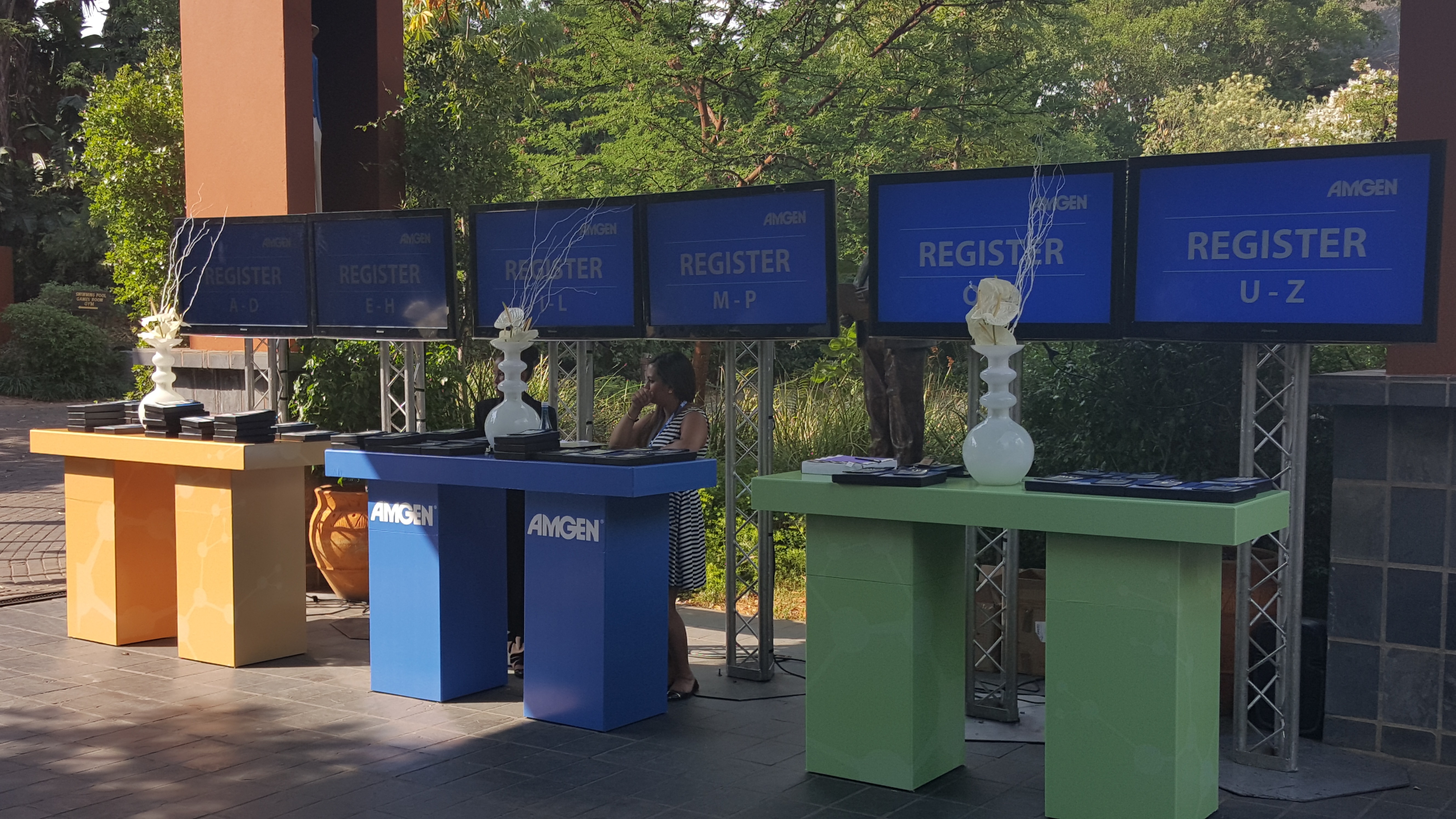 Outdoor register area with colour-coded desks in orange, blue and green, AMGEN branding, white vases, and REGISTER A–Z screens