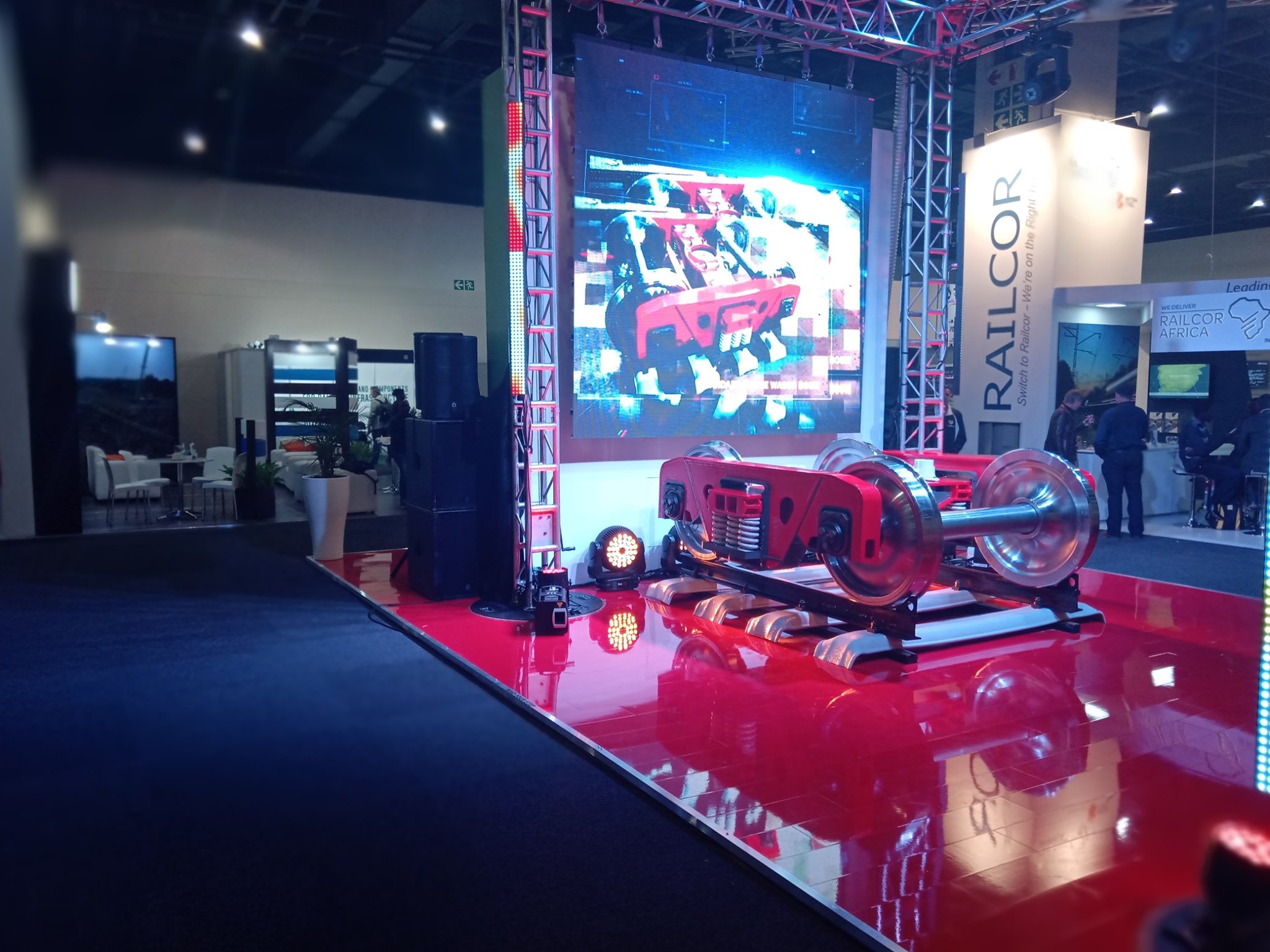Red and silver rail bogie on a glossy red platform with the RAILCOR pylon, LED wall on truss, and PA stack