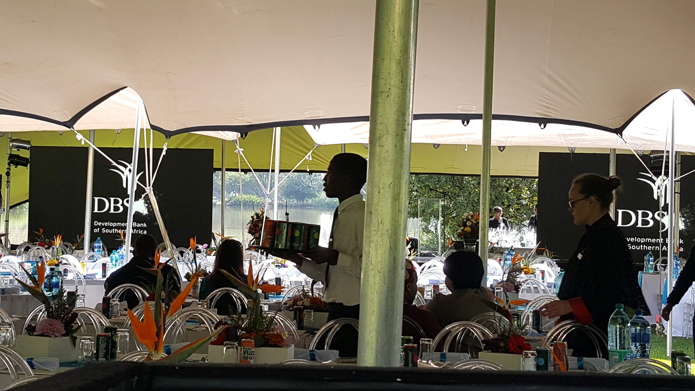 Stretch tent interior with DBSA screens, a waiter carrying a tray of drinks between banquet tables with teal runners and ghost chairs